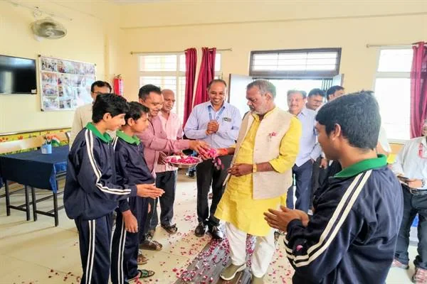 विद्यालय कक्ष में यूनिफॉर्म पहने विद्यार्थी मंत्री को फूलों की तश्तरी थमाते हुए